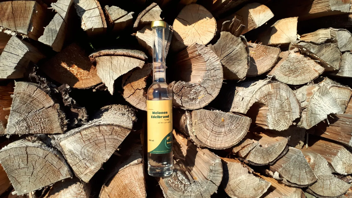 Edle Brände. Echter Genuss. Melonen Edelbrand vor gestapeltem Holz
